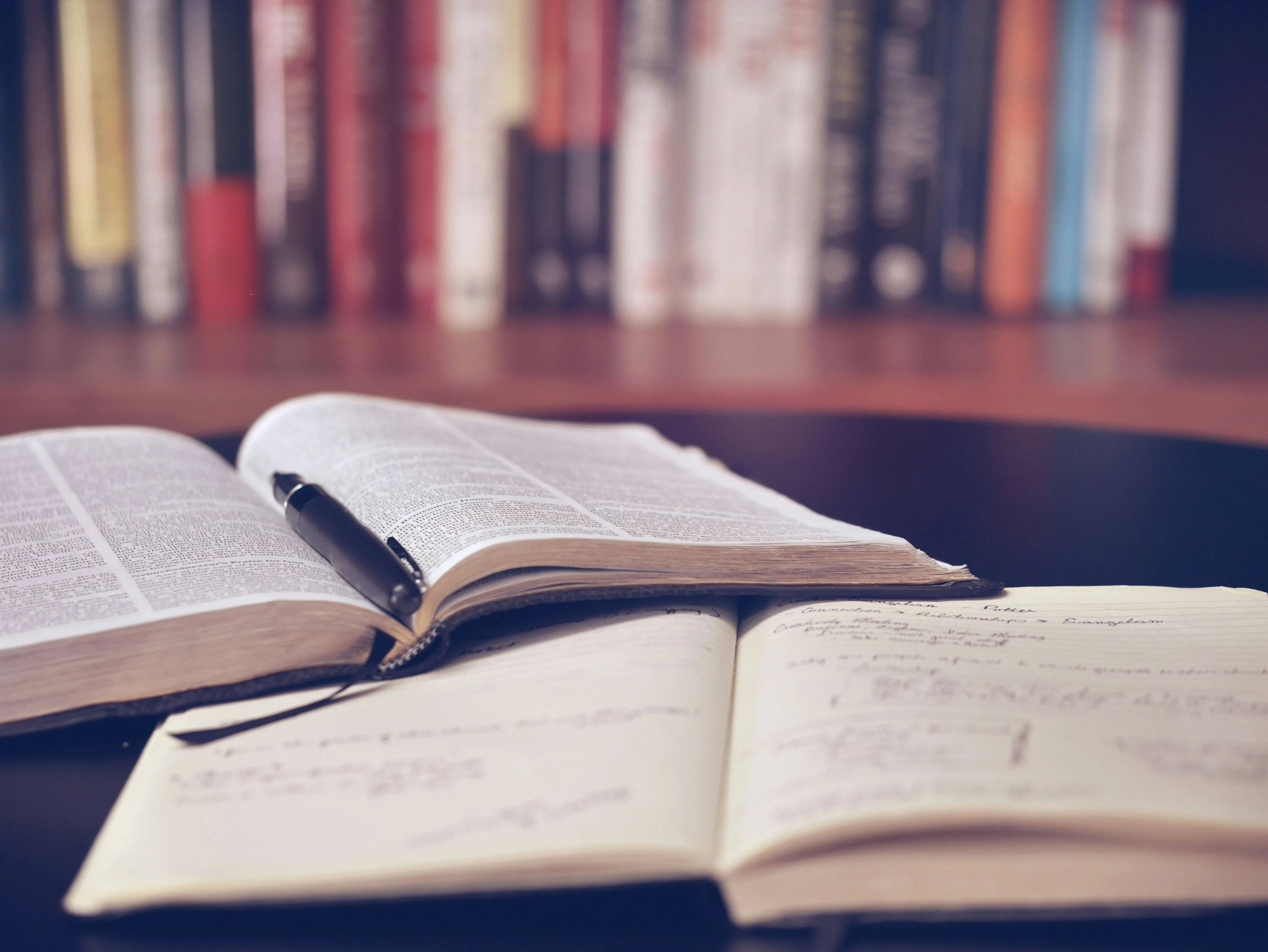 An open dictionary and a handwritten notebook with a pen, in front of a shelf of books.
